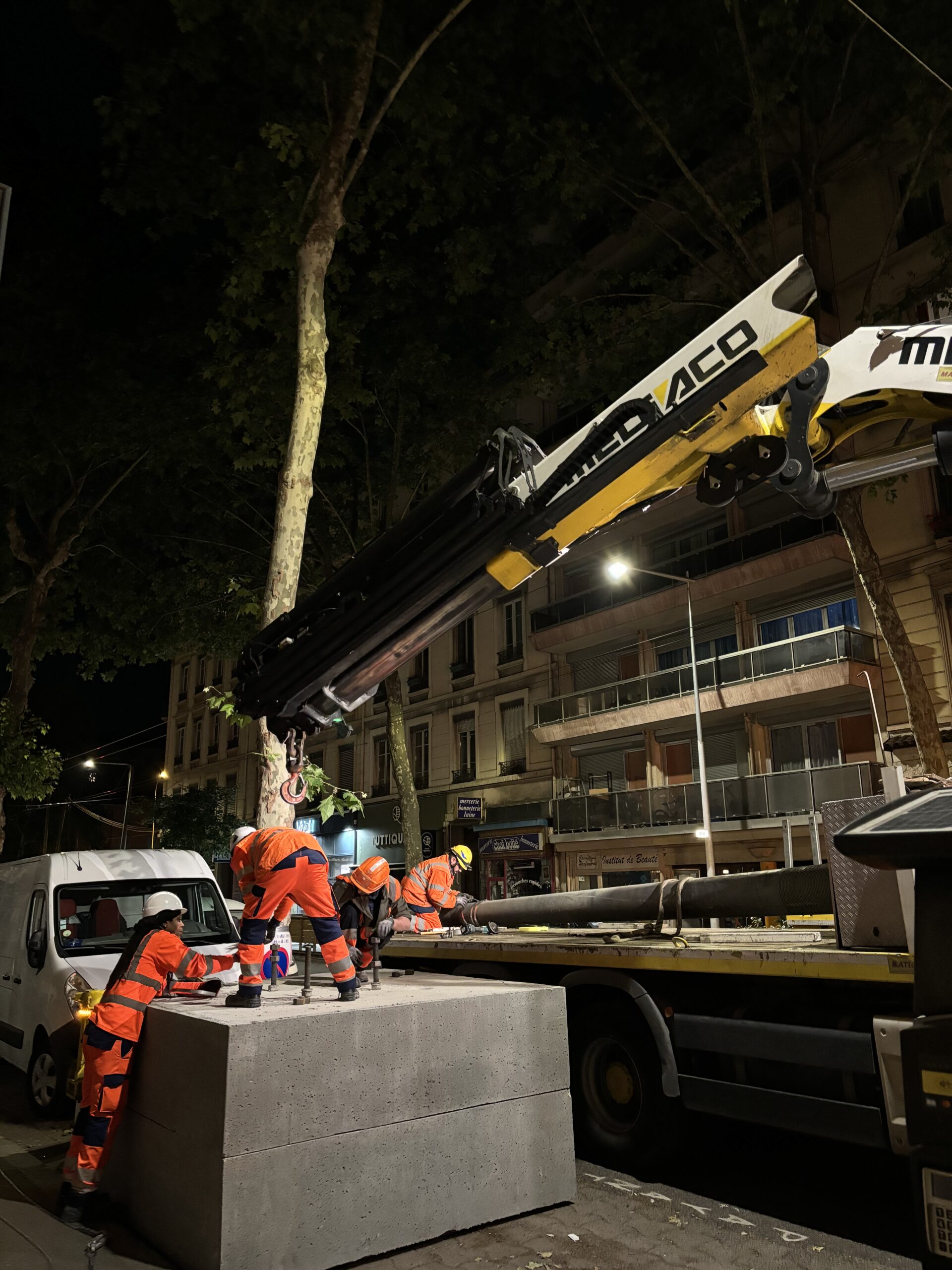 Des ouvriers portant des gilets de sécurité orange et des casques opèrent des machines lourdes la nuit. Une grue soulève des blocs de béton sur un camion. Ils se trouvent dans une zone urbaine, avec des bâtiments et des arbres visibles en arrière-plan.